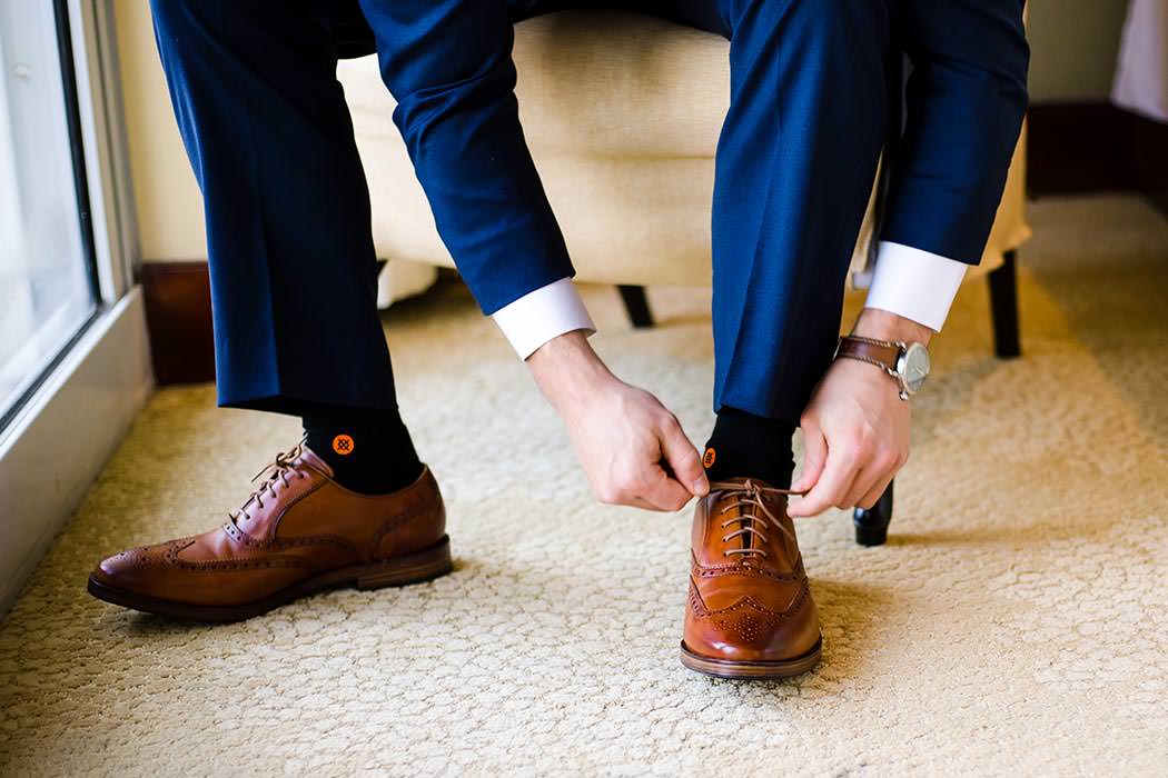 modern jewish groom putting on brown wedding shoes with navy suit