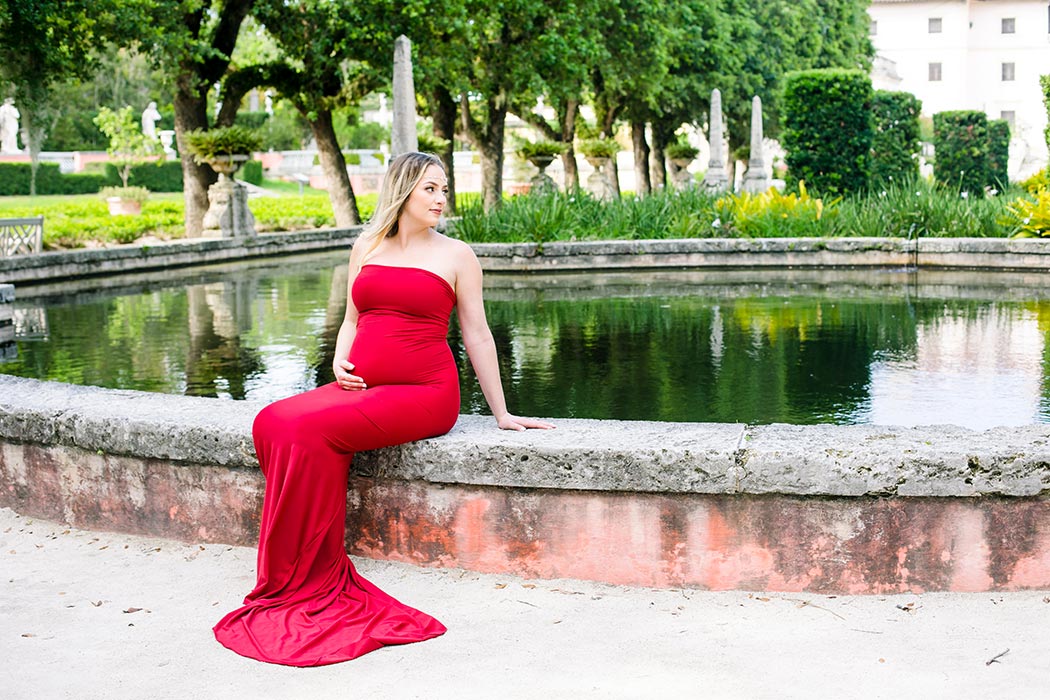 expectant mother poses in long red maternity dress at vizcaya elegant long red maternity dress | maternity photoshoot vizcaya museum miami | fort lauderdale maternity photographer