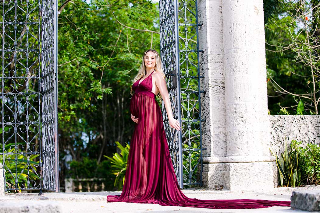 pregnant mother poses next to iron gate for vizcaya maternity photo shoot long red elegant maternity dress | maternity photoshoot posing ideas with gate | vizcaya maternity session