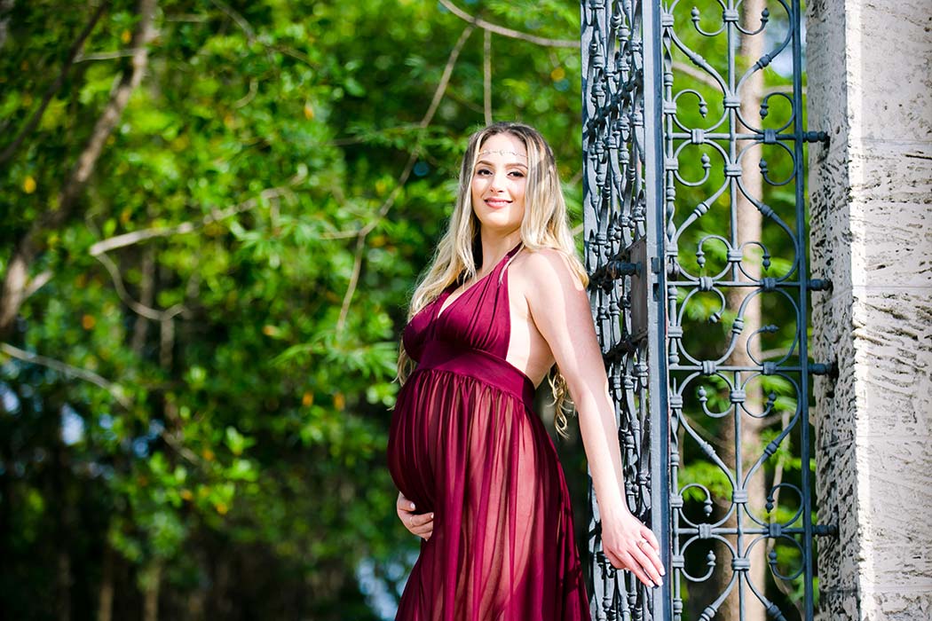 pregnant mother poses at iron gate for vizcaya maternity photoshoot unique maternity pose with long red dress at iron gates | maternity photoshoot vizcaya